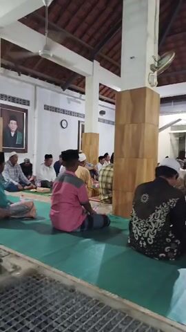 The video shows a group of men sitting on the floor in what appears to be a mosque or a similar religious gathering space. The men are dressed in traditional Islamic attire, including kufis and robes. They are seated on prayer mats, facing the same direction, suggesting they are participating in a religious service or activity. Some men are in the foreground, facing away from the camera, while others are further back, partially visible. The environment is indoors, with a tiled roof and white pillars supporting the ceiling. Lighting appears to be artificial, with ceiling lights illuminating the space. A ceiling fan is visible in the upper left corner of the frame. The atmosphere seems calm and contemplative.