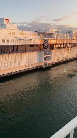 A large white ship is docked in the water, bathed in the warm light of the setting sun. The ship has multiple decks and numerous windows, many of which reflect the golden sky. There are white railings in the foreground, suggesting the video is filmed from another vessel or dock. The water is a deep green and appears calm, with a few small birds visible on the surface. The sky is a soft blend of orange, pink, and blue hues, with scattered clouds. A triangular logo, colored red and blue, is visible on the top of the ship.