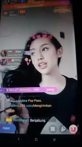 The video shows a young woman with long black hair, wearing a black top, and adorned with a filter of pink hearts around her head. She is looking to the right of the camera and appears to be speaking. The video interface indicates a live stream with various elements such as user names, chat messages, and interactive buttons like "Halo!" and "Ikuti". Some of the chat messages visible include user names and their actions, such as sending gifts or joining. The environment appears to be indoors, with a neutral background. The overall impression is a segment of a social media live stream.