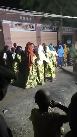 The video shows a group of people, mostly women, gathered outside a building at night. The women are wearing similar green patterned dresses. Some of them have head coverings of various colors. They are standing in a circle, moving slightly to the music. The ground is uneven and littered with small debris. In the foreground, the backs of some young people are visible, possibly watching the group. The building in the background is two stories and has a light-colored facade with dark-framed windows. The lighting is poor, suggesting it is nighttime with minimal illumination.