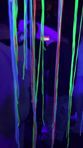 The video shows several people standing in a dark outdoor environment illuminated by blacklights, causing neon strings of blue, green, and red to glow vibrantly. The strings are hanging down from a circular structure above, creating a colorful curtain. Some individuals are partially visible through the strings, appearing to be interacting or observing the setup. The atmosphere suggests a nighttime event or gathering with decorative lighting.
