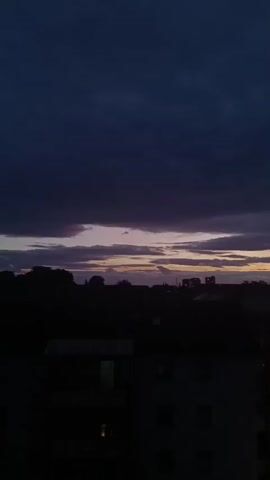 A low-angle shot captures a dark sky with dramatic clouds and a faint sunset visible on the horizon. Silhouetted against the sky are buildings and structures, some with visible chimneys. In the foreground, the dark shapes of more buildings are discernible, with a few lit windows. A white van parked on a street is illuminated by its headlights. The overall atmosphere is twilight and somewhat ominous.