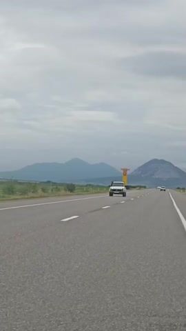 A white pickup truck drives down an empty highway. Mountains are visible in the distance. A tall yellow sign with a red stripe is on the side of the road. The sky is overcast and cloudy. The road has white lane markings.