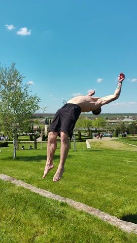 This video captures an outdoor scene at what appears to be a park or recreational area in Neofit Rilski, Bulgaria, under bright daylight conditions.

The primary subject is a man, shirtless and wearing black shorts, performing an acrobatic backflip on a well-maintained green grassy lawn. The video begins with him mid-air, upside down, with his arms extended as he completes the rotation. He successfully lands squarely on his feet, then takes a few steps forward, looking relaxed after the maneuver.

In the background, other people are visible; some are seated on benches, while others are standing or walking on the grass. The setting includes numerous trees, both deciduous and coniferous, and a low stone wall or border on the right side of the frame. The horizon reveals distant buildings or structures, suggesting a view over a town or countryside. The sky is clear and blue with a few scattered white clouds, indicating pleasant, sunny weather, likely late morning or afternoon. No text is visible in the video.