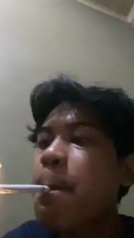 Here is a description of the video:

The video shows a close-up, low-angle shot of a young man with dark hair and a slight mustache, taking a drag from a cigarette. He is seen lighting the cigarette with a lighter. After inhaling, he exhales the smoke through his nostrils. The background appears to be a plain wall, likely indoors, with a simple ceiling visible in the top corner. The lighting in the scene is dim, enhancing the glow of the lighter and the smoke.