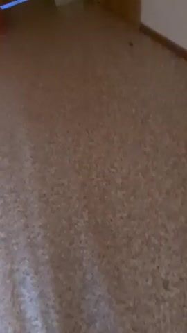 The video shows a close-up, low-angle view of a speckled, light brown floor that appears to be linoleum or a similar material. The floor has a light beige base with small, dark brown and white speckles. Along the right side of the frame, a wooden door frame is visible, with a portion of a light brown wooden door slightly ajar. The wall to the left of the door frame is a plain white. The lighting is dim, casting shadows that suggest an indoor environment, possibly a hallway or room.