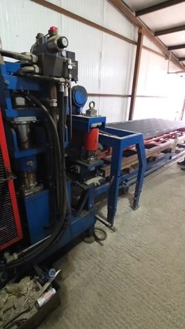 The video shows industrial machinery in what appears to be a large warehouse or factory. The primary focus is on a blue metal machine with hydraulic components and a control panel, which seems to be in operation, evidenced by sounds of machinery. Beside it, there's a metal structure, and a box filled with what looks like scrap or waste. The camera pans to show a control panel, then shifts to a broader view, revealing a long, enclosed structure filled with spherical objects, possibly metal balls or similar industrial components. The environment suggests a production or manufacturing setting. The building itself is constructed with metal beams and corrugated walls, typical of industrial buildings.