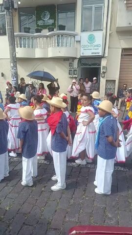 A group of young children, perhaps around eight years old, are kneeling or standing on a cobblestone street. Most of the girls wear white dresses with red and yellow accents, while most of the boys wear dark blue shirts with white polka dots and white pants. All wear hats. They appear to be performing a dance. Behind them and to the sides are a variety of adults watching, taking pictures, and generally observing. A building is in the background with a dental office called "Odontomed". There is some drizzle in the air.