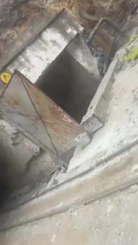 The video starts with a downward view into a dark, somewhat concealed opening. A metal door with an "X" design is partially covering the entrance. The surrounding area appears rugged with stone and rubble. The camera then abruptly switches to an exterior shot from a moving vehicle. A roadside sign and a utility box are visible. The camera is then fixed on the view through the car's windshield, showing a road leading into a town surrounded by mountains. The sky is overcast and there are a few cars on the road. Buildings and trees are visible along the roadside.