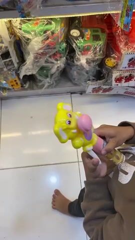 The video shows a close-up view of a child holding a yellow elephant-shaped toy with flashing lights. The toy appears to be a bubble gun, with a pink bubble wand attached to the elephant's trunk. The child's hands are visible, and they are wearing a brown long-sleeved shirt. In the background, shelves are filled with various toys, mostly packaged in plastic. Some of the toys visible include toy trucks, cars, and boxes labeled "Mystery Box." The floor is tiled with white and light gray squares. The lighting in the store is bright and artificial. The child's bare left foot is visible on the floor. The camera pans slightly to the right, revealing more shelves of toys.