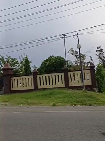 The video shows a moving vehicle's perspective, capturing the exterior of a building complex. The main entrance features a dark metal gate flanked by brick pillars topped with decorative finials. A beige fence with dark brown trim runs along the perimeter. Behind the fence, palm trees and other foliage are visible. Further down the fence, a sign with white text on a dark background reads "KEPOLISIAN NEGARA REPUBLIK INDONESIA DAERAH XXXX KEPULAUAN RIA" and "POLRI UNIT MASYARAKAT". Another section of the building shows a red-roofed structure with pillars and motorcycles parked outside. In the distance, a white structure with a BSI ATM is visible. The sky is overcast.