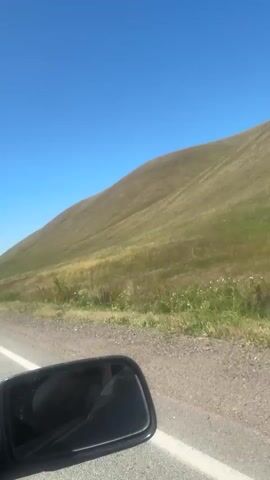 A side-view mirror of a vehicle is visible in the foreground on the left. The vehicle is on a dirt road. To the right of the road is a grassy hillside that curves upwards and to the right. The sky is clear and blue with a few small white clouds. The sun is shining. The hillside is covered in green and yellow grass. There are some small white flowers in the grass. The video is taken from a moving vehicle, as the scenery appears to be passing by.
