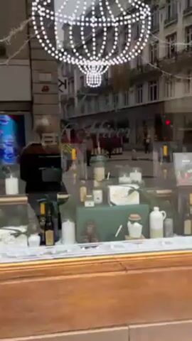 The video shows a street in a European city. The camera pans from a storefront with a decorated window display to the street scene. The store sign indicates that they personalize items. There are people walking along a wide pedestrian walkway. Buildings are on either side of the walkway and many have multiple stories. Some street decorations are visible overhead. The weather appears overcast.