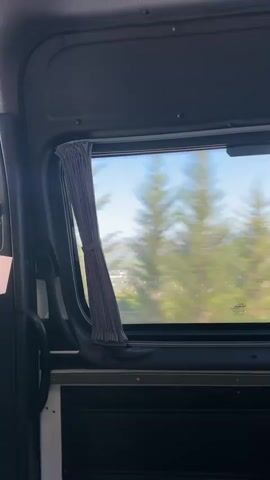 The video shows a view through a window from inside a moving vehicle, likely a bus or van. The window has a gray curtain pulled to one side, revealing a landscape outside. The landscape appears to be rural, with rolling hills, trees, and some buildings visible in the distance. The trees and landscape are blurred due to the motion of the vehicle, creating a sense of speed. The sky is visible and appears to be clear and blue. The interior of the vehicle is dark and out of focus, with the window and the landscape being the primary subject.
