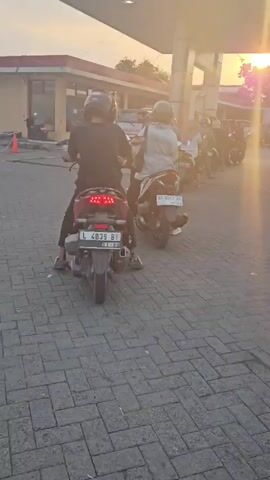 The video shows a line of motorcycles at a gas station. The sun is setting, casting a warm glow over the scene. The motorcycles are waiting to be refueled. The gas station appears to be in a rural or suburban area. The license plates of the motorcycles are visible, suggesting they are from Indonesia.