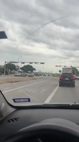 The video shows a view from the driver’s seat of a car stopped at a busy intersection under a cloudy sky. Several cars and a large Sysco truck are visible in the other lanes. A man wearing a black shirt and red shorts is walking along the curb on the right side of the road. He pauses on the corner and raises his arms up as if in celebration or excitement.