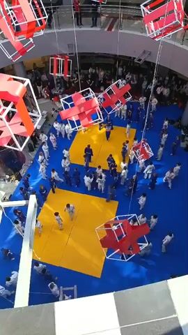 From a high angle, a large group of young people in judo uniforms are practicing on two yellow mats placed on a blue floor. There are also people in the background watching from the upper level. Red-and-white geometric decorations are suspended from the ceiling.