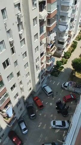 A high-angle drone shot pans over an apartment building courtyard filled with parked cars. A white truck with a loaded bed is parked on the right side of the frame. Several cars are parked haphazardly, some with doors open. The courtyard is paved with concrete and surrounded by apartment buildings. Green trees and bushes are visible on the right side of the frame. Laundry is hanging from some of the apartment balconies. Two individuals are visible in the courtyard, walking between parked cars. The overall scene appears to be a residential area on a sunny day.