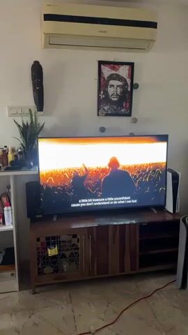 The video opens on a living room featuring a TV playing a scene with subtitles, mounted on a wooden cabinet with a Playstation console beside it. A speaker sits to the left of the TV. Above, there's an air conditioning unit and a framed portrait of Che Guevara. A decorative wooden mask is hung on the wall as well as a plant. The camera pans to the right, revealing a cluttered shelf and a portion of the room with a distressed wood table and chairs. A reddish-brown dog is lying on the tiled floor underneath the table.