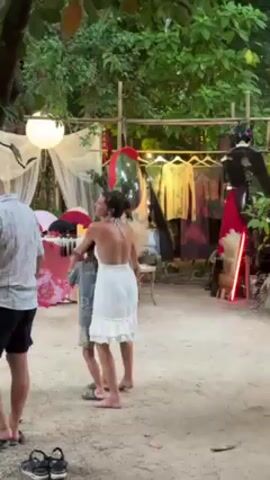 A woman in a white dress and a child are dancing outdoors on a dirt surface. To the right of them are clothing racks with various garments, and to the left, there is a display of colorful fans. A man stands to the left, observing. The area is decorated with lanterns and lights, and lush green trees surround the scene.