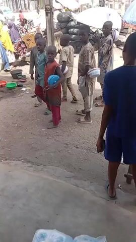 The video shows several young boys walking down a dirt road, each carrying a plastic bowl. They appear to be in a market area or a busy street with shops and stalls visible in the background. A young man is standing by a cooler, possibly selling drinks. Later in the video, two other young men are shown sitting on the ground eating from bowls, while a third watches them. They appear to be enjoying a meal together outdoors. The environment seems urban and somewhat cluttered, with scattered trash and various objects on the ground.