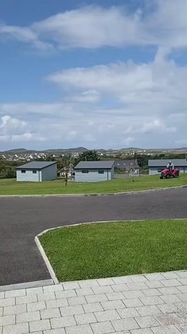 A person is riding a red tractor on a paved road next to a grassy area with several small, light blue buildings with dark gray roofs in the background. The sky is blue with white clouds. The person on the tractor is wearing a light-colored shirt and dark pants. The tractor appears to be a riding lawnmower. The scene is an outdoor setting, likely a park or residential area.