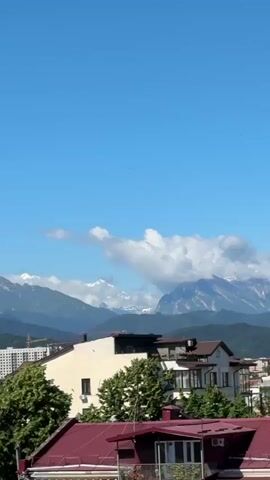 The video shows a panoramic view of a mountainous landscape with a town in the foreground. The sky is a clear blue with scattered white clouds. In the distance, several mountain ranges are visible, with the peaks of some appearing to be snow-capped. The closer mountain ranges are covered in lush green forests.

In the foreground, a town with various buildings is seen. There are multi-story residential buildings, some with balconies and windows. The roofs of some buildings are red, while others are a darker color. Trees are interspersed throughout the town. One prominent building has a blue roof. A Ferris wheel is faintly visible on a distant hilltop behind the town. The overall scene suggests a bright, sunny day in a scenic location.