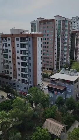 This video is a high-angle shot of an urban scene, likely taken from a balcony or window. The foreground shows a dense canopy of green trees, with a brown, shed-like structure visible at the bottom. Beyond the trees, a paved parking lot with several cars is apparent. In the mid-ground, a multi-story building with a grey facade and several balconies stands out, with cars parked in front. Further back, a cluster of modern apartment buildings of varying heights and colors dominate the landscape. Some of these buildings feature external air conditioning units and solar water heaters on their roofs. The sky above is overcast. The scene appears to be a residential area with a mix of older and newer constructions, surrounded by greenery. No people are visible in the frame.