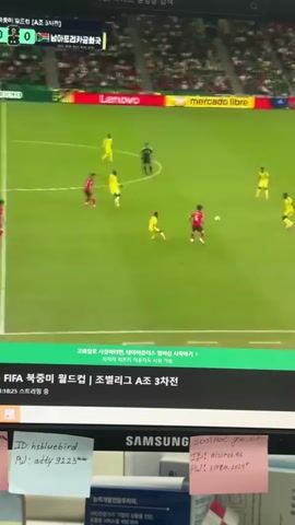 The video shows a computer monitor displaying a soccer match. On the screen, two teams are playing on a green field with spectators in the background. One team is wearing red jerseys and the other is wearing yellow jerseys. The camera focuses on a player in a red jersey dribbling the ball. In the foreground, there are sticky notes attached to the monitor, with some text written on them. One note has the text "ID: hshbluebird PW: atty 9223" written in blue ink. Another note has "soolmc.go.kr IP: oisireloke PW: korea 2024" written in pink ink. The overall scene suggests someone is watching a soccer game on their computer, with personal information logged on sticky notes.