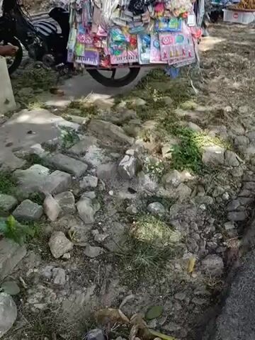 This video shows a roadside stall filled with various items, primarily small toys and accessories. The stall is mounted on what appears to be a motorcycle sidecar or a custom-built trailer, with a motorcycle parked to the left of the frame. The items on display are densely packed and include colorful hair clips, keychains, small musical keyboards, and other novelty items. The ground in front of the stall is rough and uneven, with rocks and sparse vegetation. The lighting suggests it is daytime, possibly outdoors. The video appears to be a close-up, handheld shot, with the camera moving slightly. There are no people clearly visible in the frame, though the presence of the motorcycle suggests a vendor may be nearby.