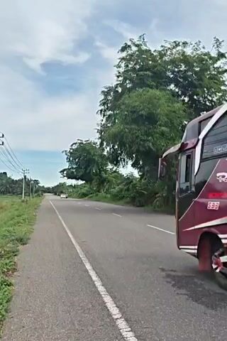 A dark red and white bus drives away from the camera on a two-lane asphalt road. The bus has a license plate on the back that says, "2244." Another car, followed by a motorcycle, drives toward the camera on the opposite side of the road. There are power lines, grass, and a field on the left side of the road. On the right, there are trees and other vegetation. The sky is partly cloudy.