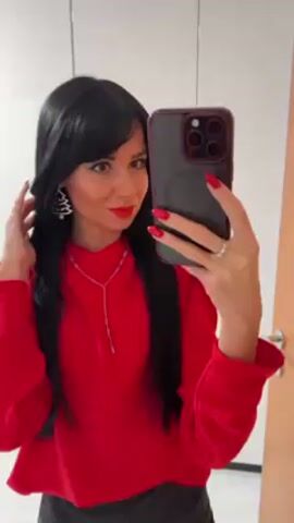 A woman takes a selfie in front of a closet with white doors and a wooden door. She wears a red long-sleeved shirt and black pants. She has black hair with bangs, red lipstick, and red nail polish. She is wearing a necklace and earrings shaped like Christmas trees. She is holding a phone with multiple cameras.
