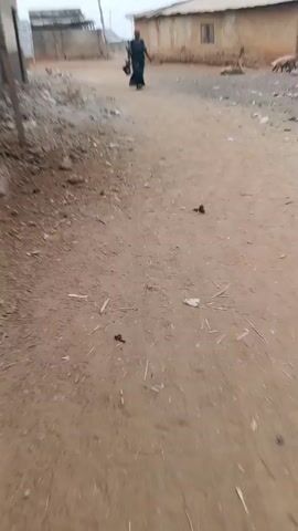 The video shows a dirt road in what appears to be a village. A woman wearing a long, dark dress is walking towards the camera, carrying a black container. In the background, there are simple, one-story buildings with pale walls and corrugated metal roofs. A few chickens and other people are visible in the distance. The ground is covered in dirt, small rocks, and scattered debris. The sky is overcast, creating a muted light.