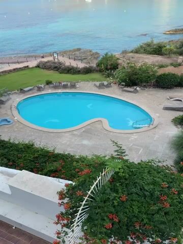 A drone shot looks down on a swimming pool with bright blue water, shaped like a kidney bean. The pool is surrounded by a paved patio with lounge chairs. Beyond the pool, a well-maintained lawn slopes down to a rocky coastline. The sea is a vibrant turquoise color, with several sailboats anchored offshore. In the background, rolling green hills dotted with sparse vegetation rise against a clear blue sky. The camera slowly pans upwards, revealing more of the bay and the distant hills.