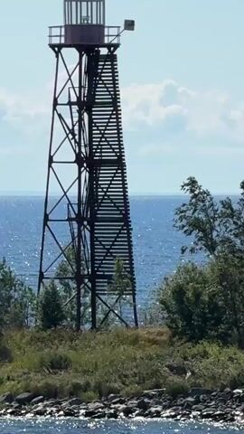 The video shows a lighthouse on a small island. The lighthouse is a tall, metal structure with a staircase winding up its side. At the top of the lighthouse is a small cabin with windows. The lighthouse is surrounded by trees and vegetation. The ocean is visible in the background, with waves gently lapping against the shore. The sky is blue and clear, with a few white clouds. The sun is shining, casting a bright light on the scene. The camera pans slowly from left to right, giving a full view of the lighthouse and its surroundings.