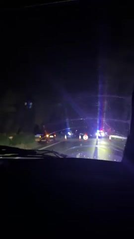 The video, filmed from inside a vehicle at night, shows a road blocked by traffic cones and a circular "no entry" sign. Police lights, predominantly blue and red, flash from vehicles ahead, illuminating the scene. The road appears wet, possibly from rain, and the surrounding environment is dark, with trees visible on the left side. The camera pans slightly, showing other vehicles and the ongoing activity on the road.