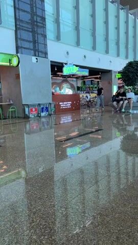The video shows the interior of what appears to be an airport terminal or a large shopping mall. The floor is highly polished and reflective, showing the ceiling and overhead lighting. On the left side of the frame, there are several large trash receptacles. In the middle ground, there is a food establishment with a bright green counter and a sign that reads "BIG BOWL". Several people are visible in and around the food establishment, including a woman in a white top and shorts walking towards the counter, a person in a black top and pants standing behind the counter, and a man and two women sitting on a bench to the right of the food establishment. Further to the right, more people are seated and standing. The overall environment is clean and modern, with large windows and a spacious feel.