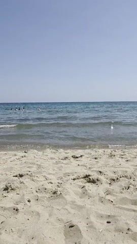 The video shows a low-angle view of a sandy beach with the ocean in the background. The sand is light brown and textured with footprints. The ocean is a clear blue with gentle waves reaching the shore. In the distance, several people are visible in the water, appearing to be swimming or wading. The sky is a clear, pale blue, indicating a sunny day. The overall impression is a peaceful and serene beach scene.