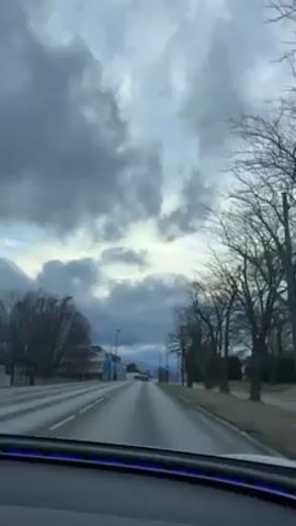 A car is driving down a road on a cloudy day. The road is paved and has two lanes. There are trees on the side of the road, and streetlights are spaced along the road. Buildings are visible in the distance. The sky is filled with dark, heavy clouds. The view is from inside the car, looking out through the windshield. A blue light is visible along the bottom of the windshield, presumably interior lighting in the vehicle. There is music playing.