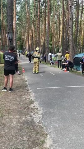 A person in a firefighter's uniform is running on a paved path through a forest. The path is lined with trees on both sides. There are spectators and tents set up along the side of the path. The person in the firefighter's uniform is running towards the camera.