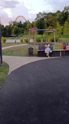 The video shows a park scene with a playground. There is a Ferris wheel in the background. In the foreground, several people are sitting on a curved bench. Two children are visible on swings. A wooden climbing structure with a rope net is also present. The ground is dark asphalt. The scene appears to be at dusk or late afternoon, with warm lighting on the playground structures.
