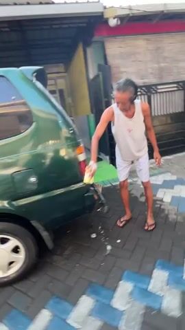 The video shows a person washing a green van in what appears to be a residential driveway. The person, wearing a white tank top and white shorts, is using a yellow sponge to scrub the rear of the van. There is soap suds and water on the van and the ground. The driveway is paved with interlocking bricks in a pattern of blue, grey, and white. In the background, there is a building with a black gate and a security camera. The sun is shining brightly, creating lens flare in the upper part of the frame.