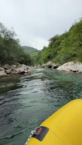This video shows a first-person perspective of a whitewater rafting trip on a river in Džajići, Bosnia and Herzegovina. The scene is set in a lush, green canyon with steep, tree-covered slopes on either side of a clear, turquoise river. Large, grey rocks line the riverbanks, and the water itself appears shallow in places, with ripples and currents indicating its movement.

The primary object visible in the foreground is the bright yellow inflatable raft, with a rope attached to its side. A person's foot, wearing a pink shoe, is visible at the bottom right of the frame, suggesting they are a participant in the rafting activity. The sky is overcast, suggesting a cloudy or perhaps misty day, which contributes to the natural and somewhat dramatic ambiance of the canyon.

The activity taking place is river rafting, a recreational outdoor activity that uses an inflatable raft to navigate a river. The movement of the raft and the perspective of the camera indicate that the raft is in motion, presumably being guided downstream. The overall mood conveyed is one of adventure and immersion in nature. There is no visible text in the media file.