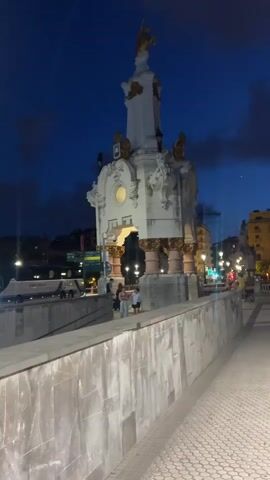 The video shows a nighttime scene of a monument on a bridge. The monument is white with gold accents and has a statue on top. There are lights illuminating the monument and the surrounding area, including streetlights and lights from buildings. A few people are visible walking on the bridge near the monument. There are also cars and a bus visible in the background. The sky is dark blue with some clouds. The video pans slightly from left to right, showing more of the bridge and the monument.