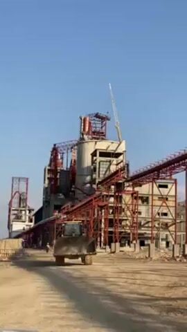 The video shows a large industrial plant under construction against a clear blue sky. The plant consists of concrete structures and red steel frameworks, including conveyor belt systems. A yellow bulldozer with a dirty blade is driving on a dirt road in the foreground, heading directly towards the viewer. To the left of the plant are some containers. In the distance, there appears to be a small hill or mountain.
