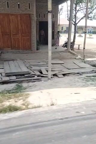 The video, captured from a moving vehicle, shows a sideways view of a rural road. On the left side of the road, there is a weathered yellow utility box attached to a pole, adorned with various posters. Further down the road, a white building with multiple windows is visible, with a bicycle mounted on its exterior wall. Adjacent to this building, a blue tarp is present amidst overgrown vegetation. The road itself appears to be asphalt, with tire tracks visible. The environment is characterized by greenery, suggesting a somewhat undeveloped area with scattered structures. The sky is visible in the upper part of the frame, appearing overcast.