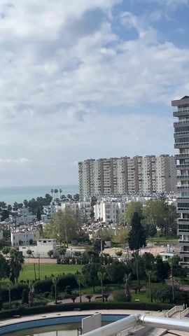 The video presents a static, medium-shot view of a coastal cityscape captured under a cloudy sky. In the foreground, a balcony railing suggests the perspective is from an elevated position. Below, a manicured park featuring palm trees and neatly arranged shrubbery extends towards a cluster of low-rise, white-roofed buildings. Beyond them, a large, high-rise apartment complex dominates the horizon. In the far distance, the turquoise sea meets the sky, completing the scene. The absence of visible people or movement contributes to a still and tranquil atmosphere.