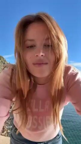 A woman with light brown hair, wearing a light pink sweatshirt with "IMPOSSIBLE" in white lettering, and blue jeans stands outside on a sunny day. She is standing on a concrete structure with a metal fence, and behind her is the ocean and a green, mountainous coastline. She initially looks down at the camera, then brushes her hair back, and spreads her arms out to the sides, balancing against the fence. The sky is blue and clear.