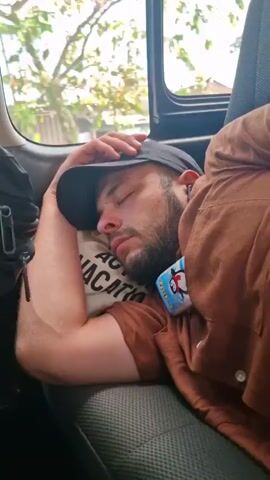 A man is asleep in the back seat of a vehicle. He has a beard and is wearing a black baseball cap, a brown t-shirt, and a brown button-down shirt. He is resting his head on a small white pillow with "VACATION" printed on it. He is also wearing an earpiece. A package of tissues with a penguin on it is clipped to his shirt. To his left is a black backpack. Outside the window, trees and buildings can be seen passing by. The vehicle appears to be moving. A sign for "Alfamart" is visible in the background.
