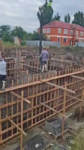 The video shows a construction site with several workers. In the foreground, a man in a blue shirt and white boots is operating a concrete pump hose. He appears to be directing the flow of concrete into a formwork structure. Behind him, another worker in a brown shirt is visible. The area is filled with wooden formwork, rebar, and various construction materials. In the background, a large red building with white trim stands under a cloudy sky. Trees and other vegetation are also present. The overall scene depicts the process of pouring concrete for a foundation.